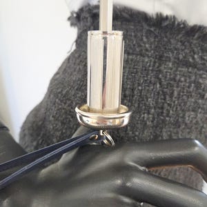 Pu&ograve; includere: Un tappo per bottiglia argentato, cilindrico, con scanalature verticali e una piccola base rotonda. Un cinturino in pelle blu scuro &egrave; attaccato al tappo. Lo sfondo comprende un tessuto grigio e una camicia bianca.