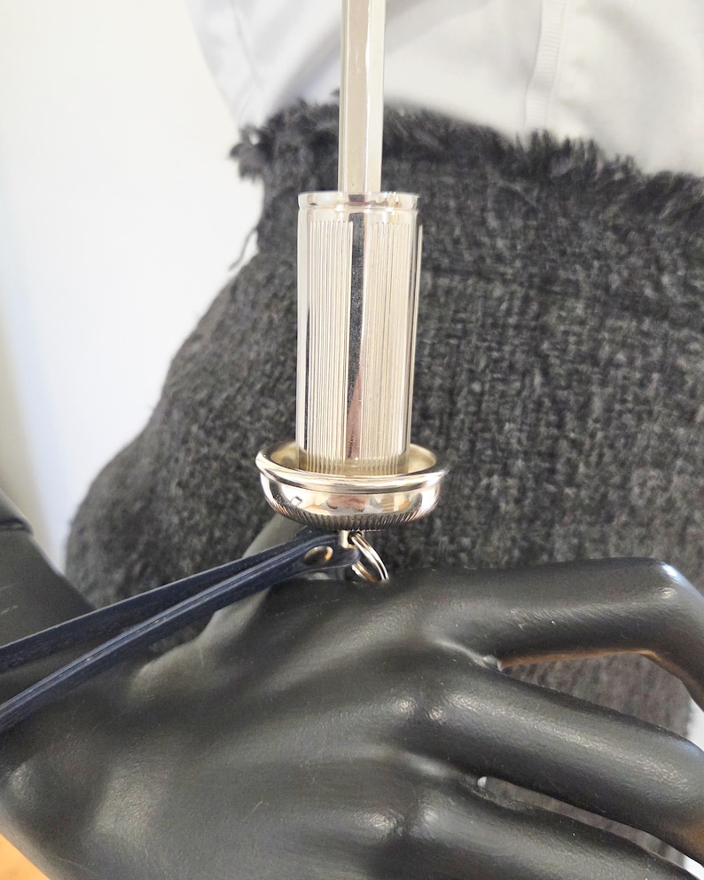 Pu&ograve; includere: Un tappo per bottiglia argentato, cilindrico, con scanalature verticali e una piccola base rotonda. Un cinturino in pelle blu scuro &egrave; attaccato al tappo. Lo sfondo comprende un tessuto grigio e una camicia bianca.