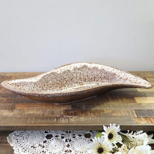 Peut inclure: Un bol décoratif en céramique marron et blanc avec une forme allongée unique. Le bol a une surface texturée avec un motif tacheté. Le bord est d'une couleur plus claire et le bol est posé sur une surface en bois.