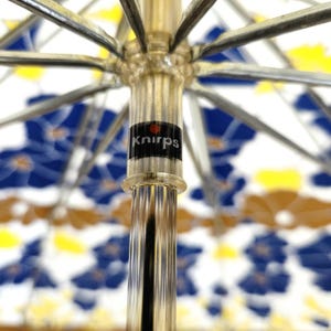 Pu&ograve; includere: Primo piano di un ombrello aperto con un motivo floreale blu, giallo e marrone su sfondo bianco. La struttura dell'ombrello &egrave; argentata e il manico trasparente con un'etichetta nera con la scritta "Knirps".