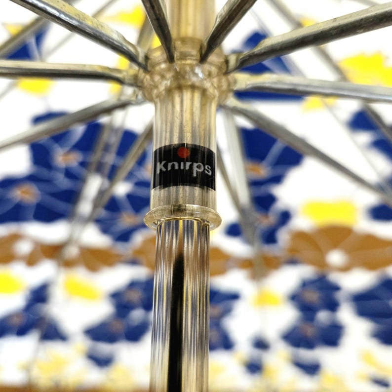 Pu&ograve; includere: Primo piano di un ombrello aperto con un motivo floreale blu, giallo e marrone su sfondo bianco. La struttura dell'ombrello &egrave; argentata e il manico trasparente con un'etichetta nera con la scritta "Knirps".