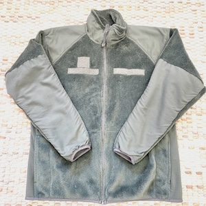 May include: A gray fleece jacket with a full-length zipper and a high collar. The jacket has reinforced panels on the shoulders and sleeves. There are two rectangular patches on the chest. The jacket is laid flat on a textured surface.