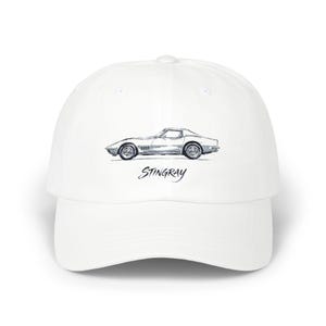 Może przedstawiać: Biała czapka z daszkiem z detalami czarno-białego szkicu klasycznego samochodu sportowego z profilu. Pod ilustracją samochodu znajduje się napis STINGRAY wydrukowany stylizowaną, kursywną czarną czcionką na przednim panelu czapki.