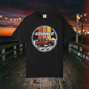 Op de afbeelding: Een zwart t-shirt met een cirkelvormige afbeelding van een rode SUV, de tekst 4RUNNER en verticale strepen in oranje, geel en crème. Het ontwerp bevat een berglandschap op de achtergrond binnen de cirkel, alles in een verweerde stijl.