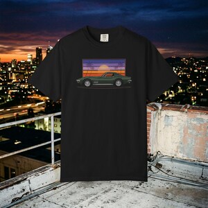 Może przedstawiać: Czarny t-shirt z krótkim rękawem z nadrukiem graficznym przedstawiającym ciemnozielony zabytkowy samochód sportowy, umieszczony na tle prostokątnego zachodu słońca z sylwetkami palm w fioletowe, pomarańczowe i żółte poziome pasy.