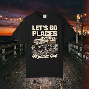 Op de afbeelding: Een zwart t-shirt met korte mouwen en een grafische print van een terreinwagen op rotsachtig terrein. De tekst boven het voertuig luidt "LET'S GO PLACES" en de tekst eronder "4Runner 4x4" in een gestileerd wit lettertype.