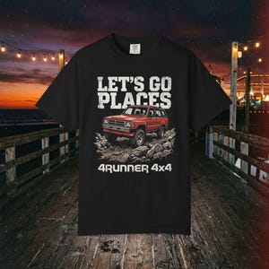 Op de afbeelding: Een zwart t-shirt met korte mouwen en een grafische print van een rode SUV op rotsachtig terrein. De tekst boven het voertuig luidt "LET'S GO PLACES" en de tekst eronder "4RUNNER 4x4" in een wit, verweerd bloklettertype.