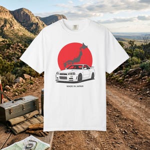 May include: A white t-shirt featuring a graphic of a white sports car positioned in front of a large red circle and a dark gray map of Japan. The text "MADE IN JAPAN" is printed in black capital letters directly below the car illustration.
