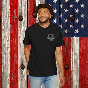 Puede incluir: Camiseta negra con un pequeño logotipo blanco en el pecho. El logotipo presenta el texto "XJ Cherokee 4x4" dentro de un diseño de escudo. El fondo es una bandera americana de madera.