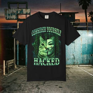 Może przedstawiać: Czarny t-shirt z nadrukiem graficznym przedstawiającym pysk kota połączony z maską Guya Fawkesa. Wzór zawiera zielony kod binarny w tle oraz napis "CONSIDER YOURSELF HACKED" wykonany pogrubioną, metaliczną zieloną czcionką.