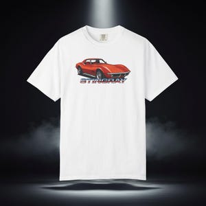 Może przedstawiać: Biały t-shirt z krótkim rękawem z grafiką przedstawiającą jaskrawopomarańczowy samochód sportowy na klatce piersiowej. Pod samochodem znajduje się napis STINGRAY wykonany pogrubioną, stylizowaną niebiesko-czerwoną czcionką. Klasyczny okrągły dekolt.