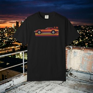 Może przedstawiać: Czarny t-shirt z krótkim rękawem z nadrukiem graficznym przedstawiającym profil zabytkowego pomarańczowego samochodu sportowego. Samochód umieszczono nad poziomymi pasami w odcieniach żółci, pomarańczu i czerwieni, z dwiema małymi palmami po prawej.