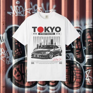 以下が含まれることがあります： 白いTシャツ。銀色のスポーツカーのグラフィックと「TOKYO STREET RACING HACHIYA」の文字。デザインには赤い円と日本語のテキストが含まれています。Tシャツはクルーネックで半袖です。