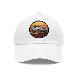 Puede incluir: Una gorra de béisbol blanca con un parche circular en la parte delantera. El parche muestra una ilustración de un SUV beige sobre un fondo de rayas horizontales naranjas, amarillas y marrones, con un borde negro cosido.