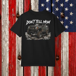 May include: A black t-shirt featuring a graphic of a dark-colored off-road vehicle driving over rocks. The text DON'T TELL MOM is printed in white above the vehicle. Two skeleton figures are visible inside the vehicle, one driving and one waving.