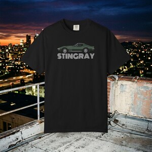 Może przedstawiać: Czarny t-shirt z krótkim rękawem z grafiką ciemnozielonego samochodu sportowego z profilu. Pod samochodem znajduje się napis STINGRAY wydrukowany białą, pogrubioną czcionką bezszeryfową z poziomymi liniami przechodzącymi przez litery.