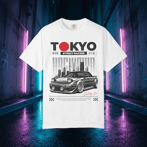 以下が含まれることがあります： 黒いスポーツカーのグラフィック、赤い円の上に「TOKYO」の文字、そして「STREET RACING」がプリントされた白いTシャツ。デザインには日本語のテキストと都市景観のシルエットが含まれています。