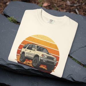 May include: Cream-colored t-shirt featuring a graphic of a beige SUV driving against a sunset backdrop of orange and yellow stripes. The SUV is kicking up dust, suggesting off-road action. The shirt is folded on a dark, textured surface.