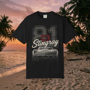 Może przedstawiać: Czarny t-shirt z krótkim rękawem z nadrukiem graficznym samochodu C3 Corvette Stingray. Projekt zawiera duży napis 81, czerwony napis C3 oraz biały napis kursywą Stingray umieszczony nad widokiem z przodu samochodu sportowego.