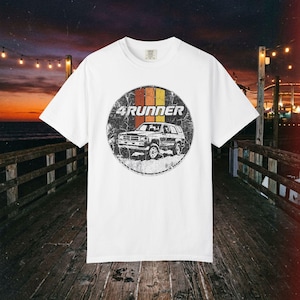 Op de afbeelding: Een wit t-shirt met korte mouwen en een ronde afbeelding op de borst. De afbeelding toont een zwart-witte illustratie van een 4Runner SUV, met daaroverheen drie verticale strepen in oranje, beige en geel, en de tekst 4RUNNER.