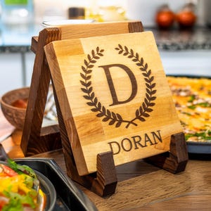 May include: A personalised wooden chopping board with a stand. The board is engraved with a laurel wreath and the letter "D" in the centre. The name "DORAN" is engraved below the wreath.