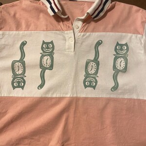 May include: Pink and white polo shirt with a cat clock design. The shirt has a white collar with navy and burgundy stripes. The cat clock design is repeated across the white horizontal stripe.