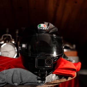 May include: A black motorcycle helmet with a clear visor and a green, white, and red sticker that says "AGV" on the top. A black action camera with the text "ACTION 5" is mounted on the front of the helmet.