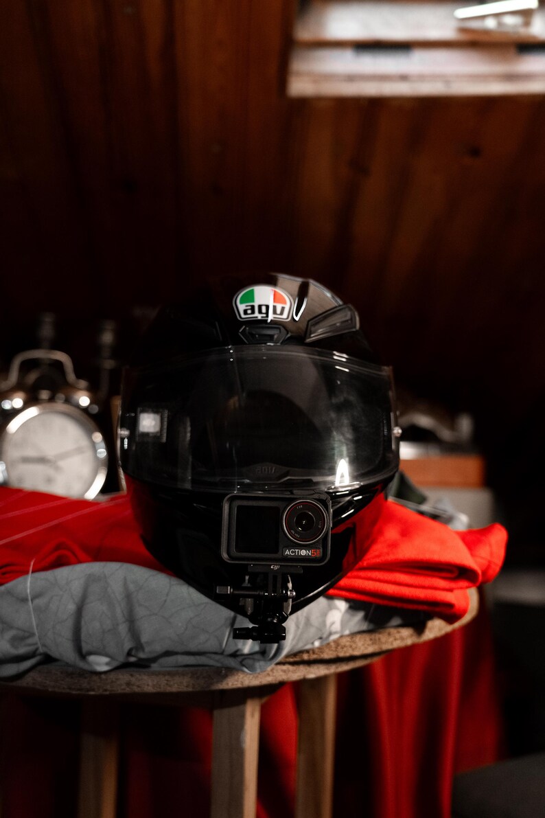 May include: A black motorcycle helmet with a clear visor and a green, white, and red sticker that says "AGV" on the top. A black action camera with the text "ACTION 5" is mounted on the front of the helmet.