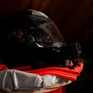 May include: A black motorcycle helmet with a clear visor and a small black action camera attached to the side. The helmet is sitting on a red and gray fabric.