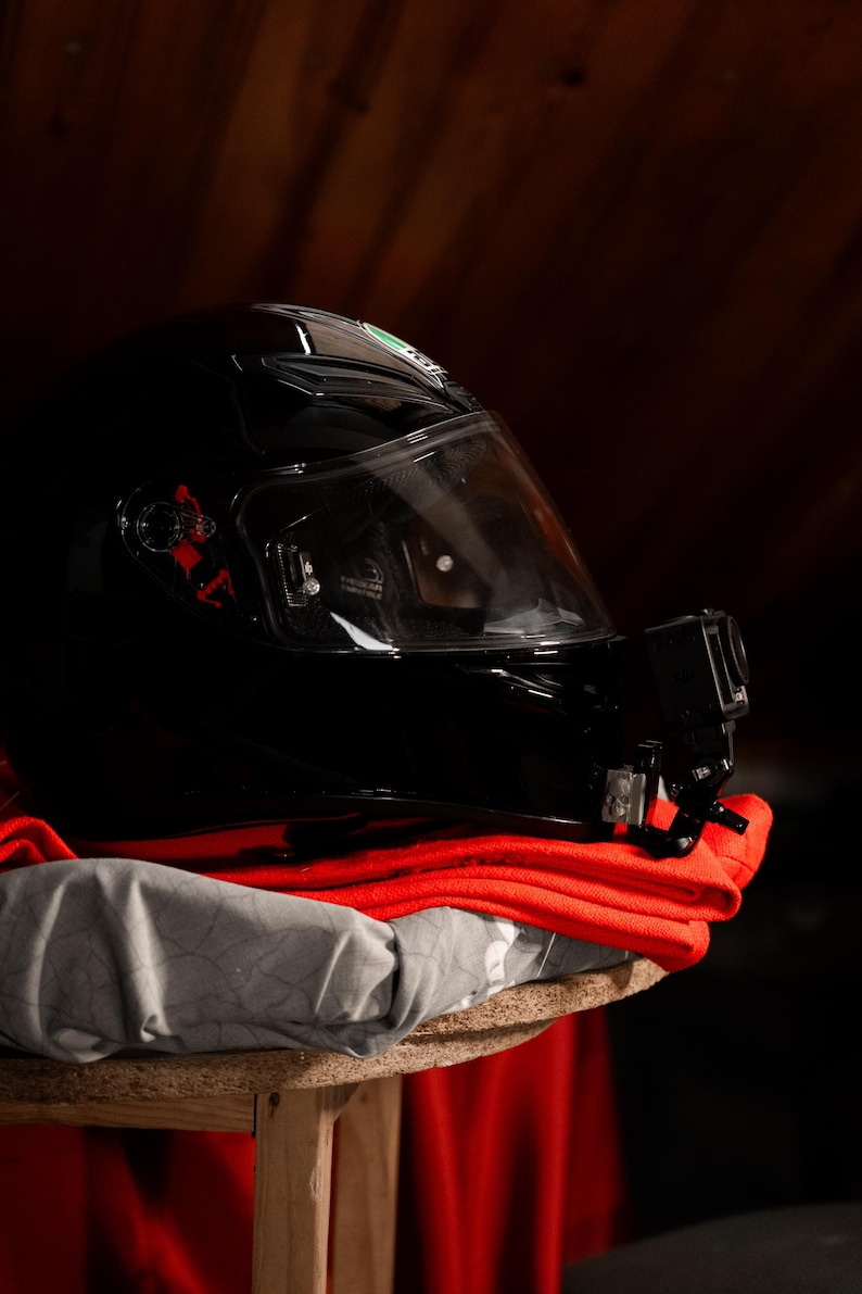 May include: A black motorcycle helmet with a clear visor and a small black action camera attached to the side. The helmet is sitting on a red and gray fabric.