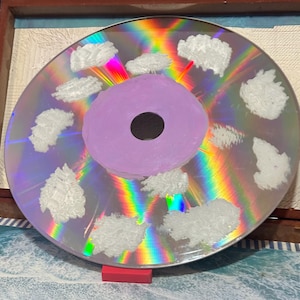 May include: A silver compact disc with a purple center and white clouds painted on the surface. The disc has a rainbow iridescent sheen.