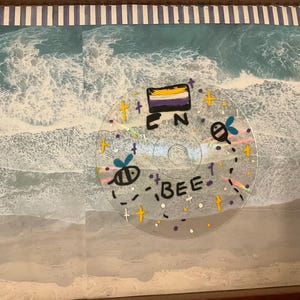 May include: A clear disc with a painted design featuring a bee, a rainbow, and a yellow, white, and purple flag. The disc also has the words "bee" and "EN" written on it.