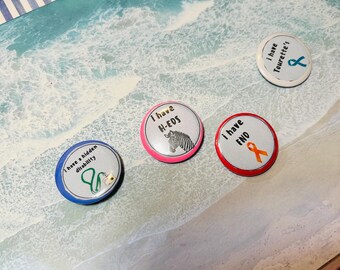 May include: Four round button badges with different awareness ribbons and text. The badges are on a wooden surface with a blue and white striped border. The text on the badges reads: "I have a hidden disability", "I have H-EDS", "I have FND", and "I have Tourette's".