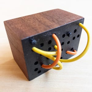 May include: A wooden cube with a dark brown top and black sides. The black sides have several small holes and are intertwined with yellow and orange flexible tubes. The cube is likely a puzzle or a decorative object.