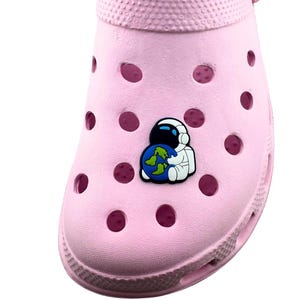 May include: A light pink clog shoe with multiple ventilation holes. The shoe features a small astronaut charm holding a blue and green globe. The shoe has the text "SIZE 5/6 ALL MAN MADE MATERIAL MADE IN CHINA" on the inside.
