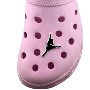 May include: A light pink clog-style shoe with multiple ventilation holes. A black silhouette of a person is affixed to the front of the shoe. The shoe is made of a rubber-like material.