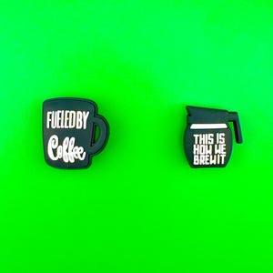 May include: Two black rubber charms with white text. One charm is a coffee cup with the text "Fueled by Coffee". The other charm is a coffee pot with the text "This is How We Brew It".