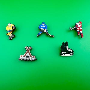 May include: Five colorful hockey-themed shoe charms. The charms include a hockey player in a yellow jersey, a hockey player in a blue jersey, a hockey player in a red jersey, a hockey stick and puck, and a black hockey skate.