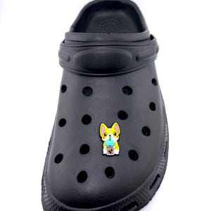 May include: Black clog shoe featuring a colourful charm of a corgi dog holding a boba tea. The charm is yellow, white, and blue. The shoe has multiple holes and a pivoting heel strap.
