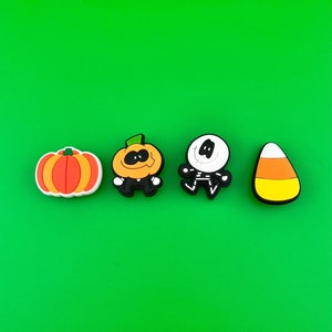 May include: Four Halloween-themed charms: an orange pumpkin, a jack-o'-lantern, a white ghost with a black skeleton, and a yellow and orange candy corn.