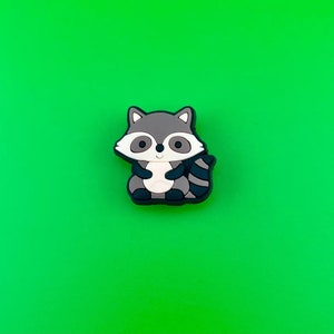 May include: A small, gray and black raccoon figurine with a white belly and a black nose. The figurine is sitting on a green background.