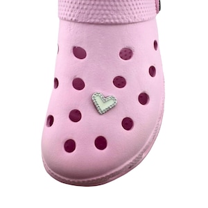 May include: A light pink clog shoe with multiple round holes. A silver heart-shaped charm with clear rhinestones is attached to the shoe. The shoe is made of a rubbery material.