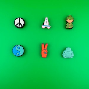 May include: Six colorful rubber shoe charms on a green background. The charms include a peace sign, a praying hands symbol, a yin yang symbol, a peace sign hand gesture, a meditating Buddha, and a meditating person with a green hat.