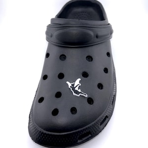 May include: Black clog shoe with a decorative charm. The shoe has multiple ventilation holes and a textured sole. The charm is a white and black design of a fairy. The shoe is made of a rubber-like material.
