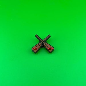 May include: A brown and black enamel pin with two crossed oars. The oars have the words "WE ROW" written on them in red.