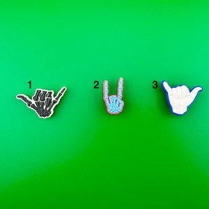 May include: Three different hand gesture charms, each with a different design. The first charm is a black skeleton hand making a shaka sign. The second charm is a light blue skeleton hand making a rock and roll sign. The third charm is a white skeleton hand making a shaka sign with a blue outline.