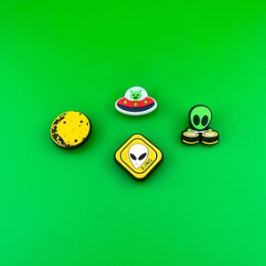 May include: Four colorful alien-themed rubber charms on a green background. The charms include a flying saucer, an alien with drums, a yellow moon, and a yellow road sign with an alien and the text "X-ING".
