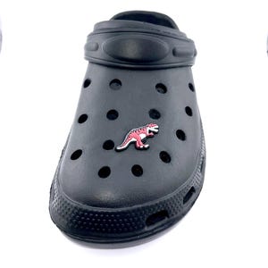 May include: Black clog shoe with a pink and white dinosaur charm. The shoe has multiple ventilation holes and a textured toe. The charm is a Tyrannosaurus Rex design.