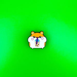 May include: A brown and white cartoon hamster with pink cheeks is drinking from a blue and white boba tea cup with a purple straw. The hamster is on a green background.