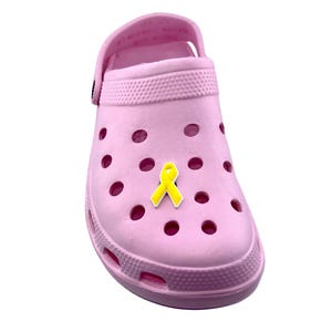 May include: A light pink clog shoe with a yellow ribbon charm. The shoe has multiple ventilation holes and a pivoting heel strap. The yellow ribbon charm is in the center of the shoe. The shoe is made of a rubber-like material.
